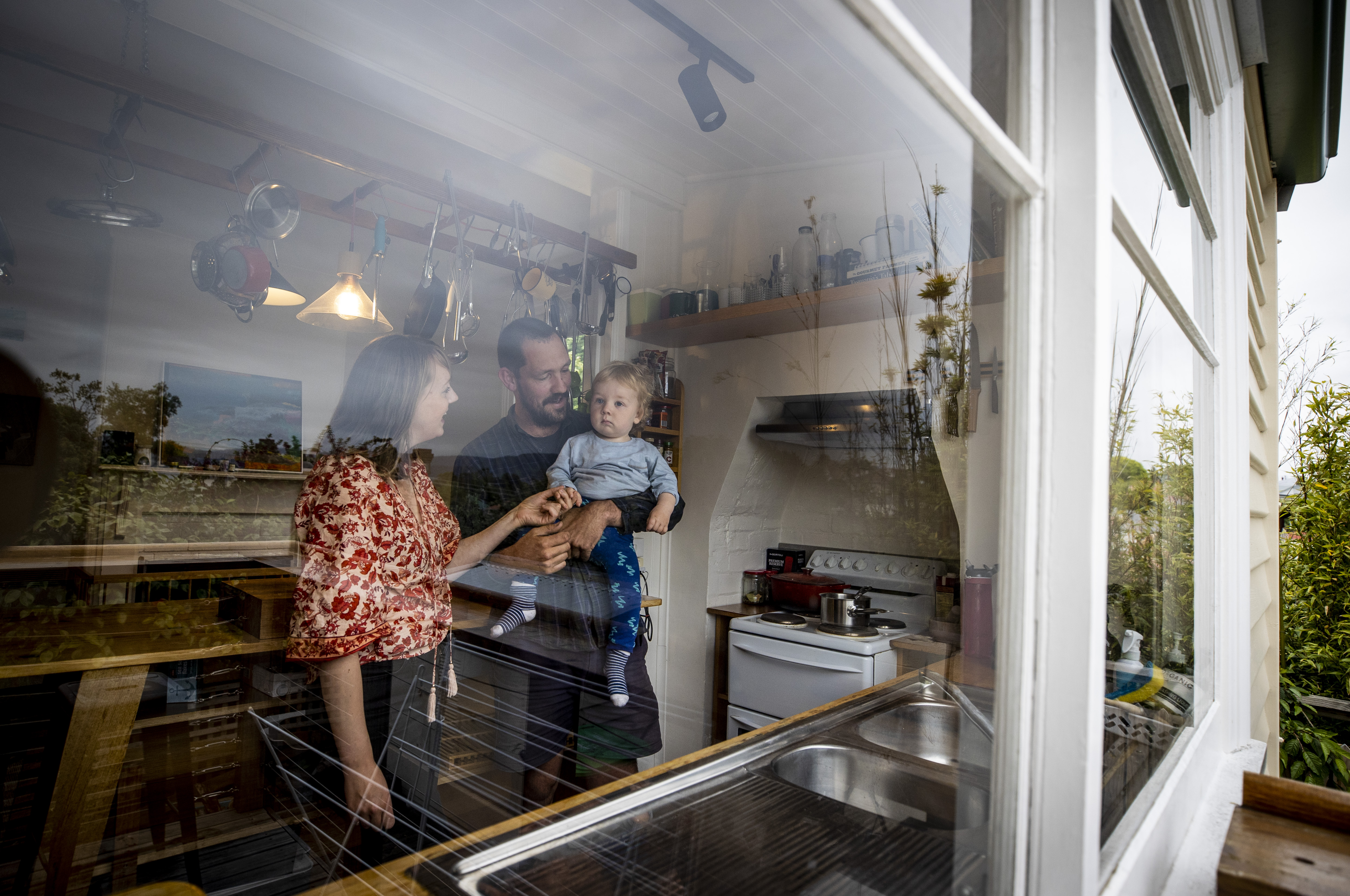 A mother and father play with their baby boy. The photo has been taken from outside through their kitchen window.